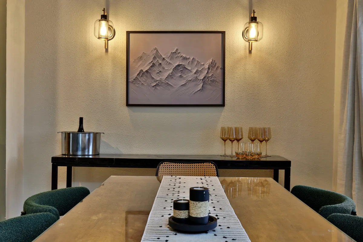 A stylish dining area featuring a wooden dining table, golden glasses set for a meal, and mountain-themed artwork on the wall. The mood is elegant yet cozy, perfect for entertaining or casual dining.