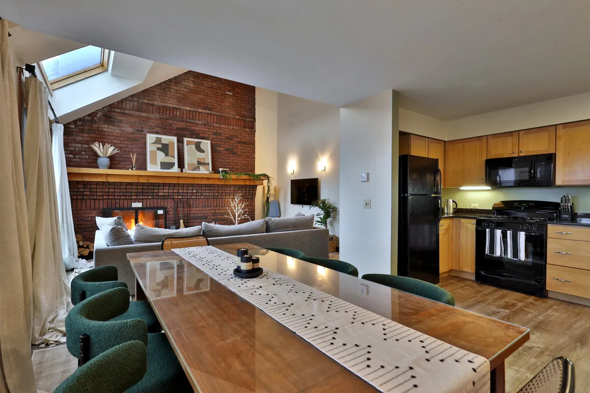 An inviting open-plan dining area with a dining table set adjacent to a sofa, exposed brick wall adorned with artwork, and a visible kitchen in the background. The scene exudes warmth and comfort, ideal for group meals and conversations.