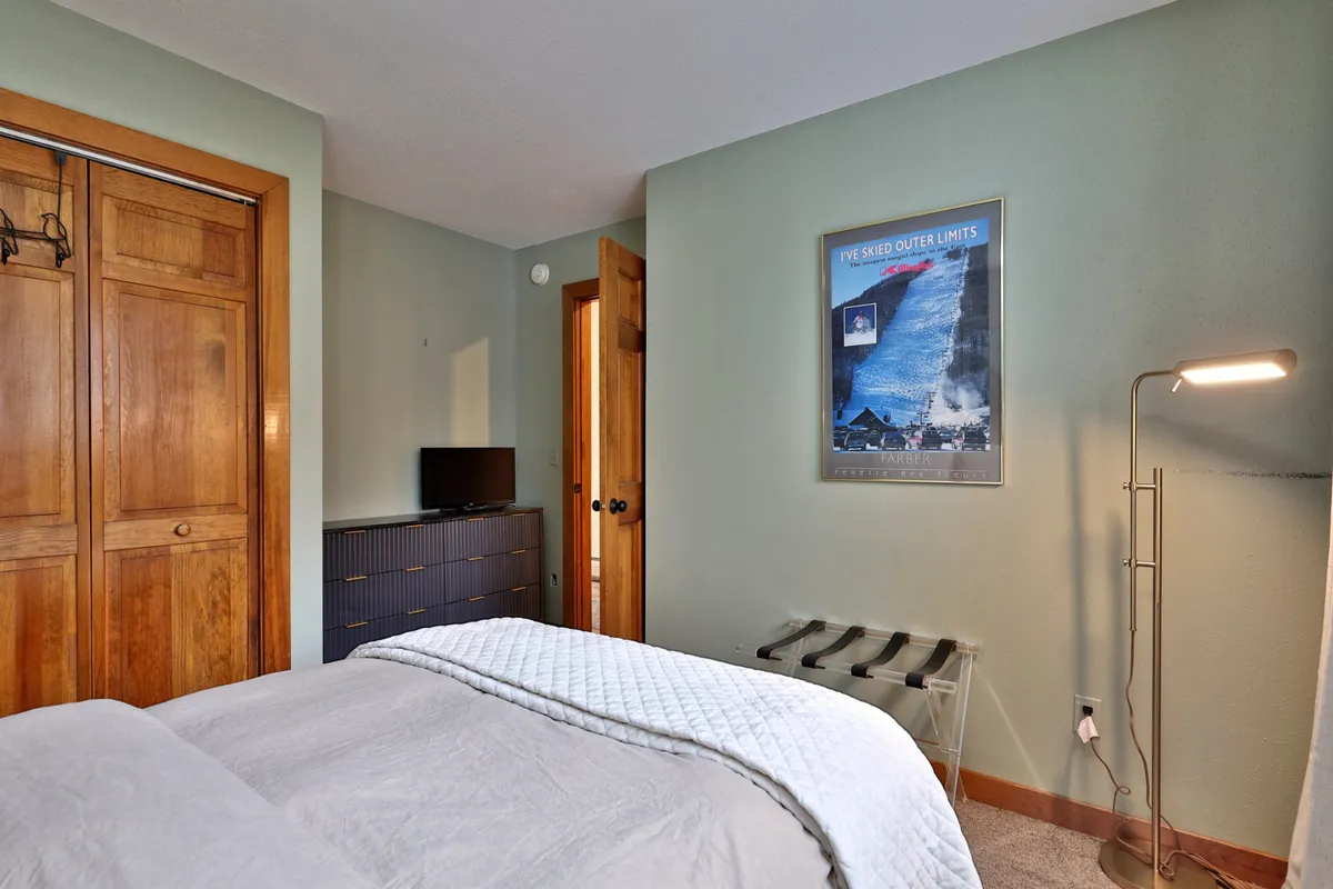 A comfortable bedroom with a queen-sized bed adorned with a quilt, framed wall art, wooden closet doors, a floor lamp, and a television. The mood is inviting, cozy, and ideal for relaxation or winding down in private.
