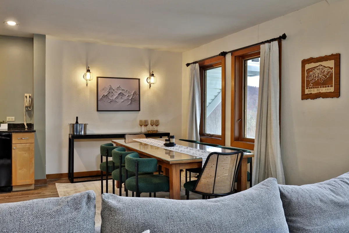A bright dining area featuring a large table with ample seating, mountain-inspired artwork on the walls, and a sofa nearby. Large windows flood the space with light and offer scenic views, setting a picturesque backdrop for gatherings.