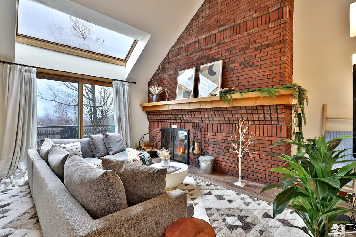 The hero image showcases a spacious, sunlit living area featuring a plush sectional sofa, a rustic brick fireplace, soaring skylights, and large windows that frame the natural Vermont landscape. The space exudes warmth and comfort, ideal for gatherings, relaxation, and après-ski moments.